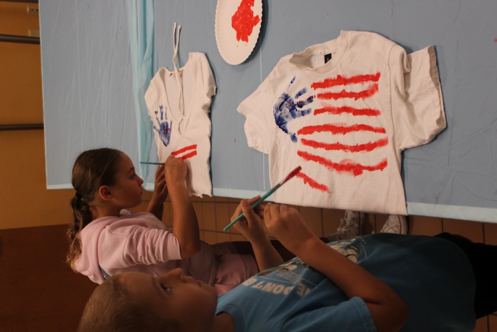 Two campers carefully paint red stripes on white T-shirts decorated with blue handprints during a 4th of July shirt-making activity at South Cape Community Center summer camp.